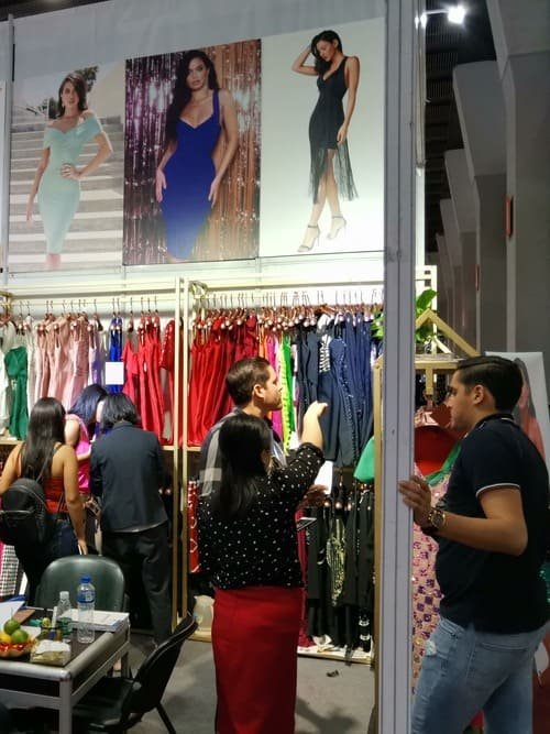 A boutique display showcases racks of colorful dresses alongside three large posters of models in evening gowns, reminiscent of a Canton Fair clothing exhibition. Shoppers browse and discuss amidst a table with water bottles and tools in the foreground.