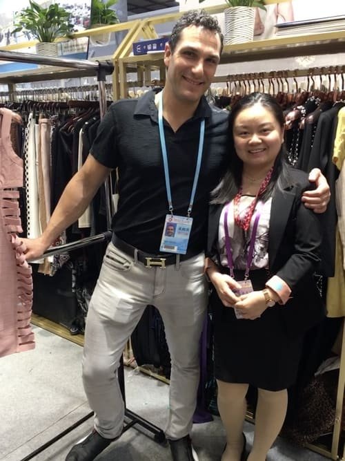 A man and a woman stand smiling in front of a clothing rack displaying various garments at the Canton Fair. The man wears a black polo shirt and light pants; the woman is dressed in a black blazer and black dress with a pink necklace, capturing the essence of this prominent clothing fair in May.