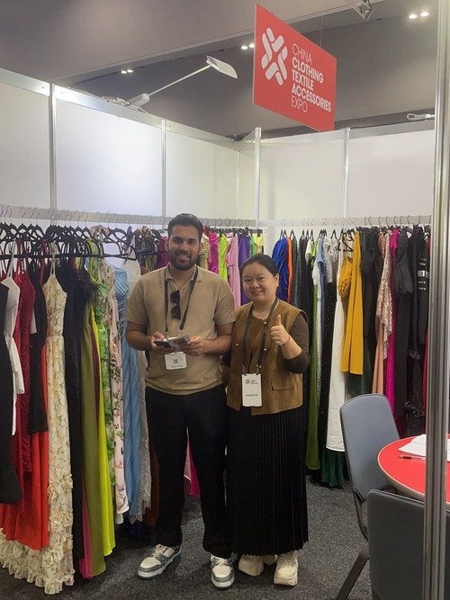 Two people stand smiling in front of a booth at the China Clothing Textile Accessories Expo, reminiscent of the vibrant energy at the Melbourne Fashion Show. They are surrounded by colorful garments on racks, with a red sign displaying the expo name above them.