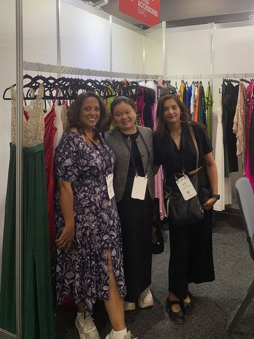Three women stand and smile for a photo at Melbournes fashion trade show. Behind them, colorful dresses fill the racks of the bustling clothing booth. Clad in business-like attire with name badges on lanyards, they embody the vibrant spirit of the event.