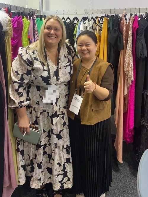 Two women smiling and posing together at the Melbourne Garment Trade Show. The woman on the left is wearing a floral dress and holding a clutch, while the woman on the right gives a thumbs up. Colorful dresses hang prominently in the background, adding to the vibrant atmosphere.