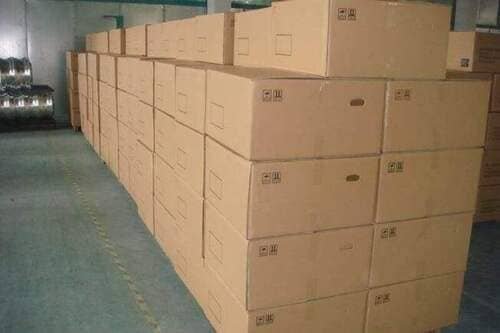 Stacks of large cardboard boxes are neatly arranged in rows inside a warehouse. The floor is smooth concrete, and the boxes are uniform in size and color, with some markings on their sides.