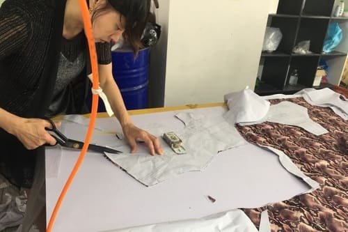 Person using scissors to cut fabric patterns on a table, with patterned and plain fabric pieces. An orange hose hangs nearby, and shelves with various items are in the background.