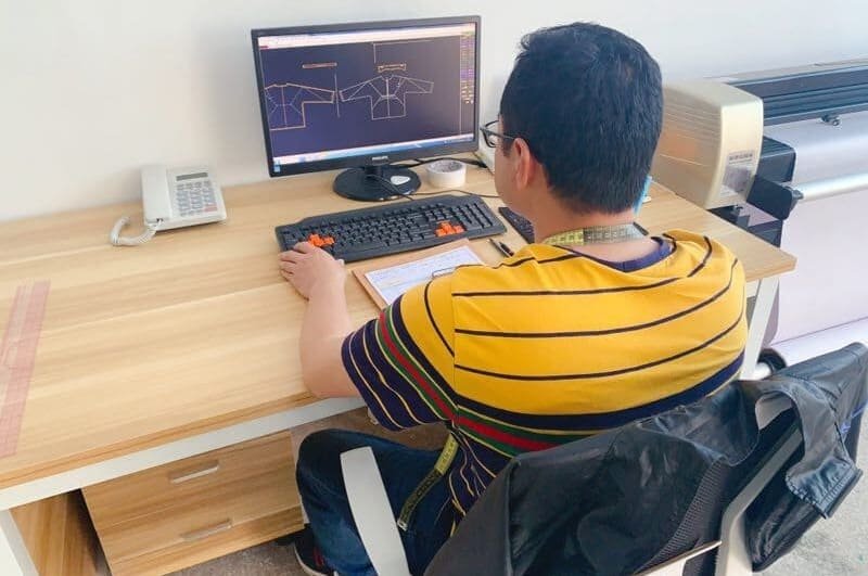 A person wearing a yellow striped shirt is sitting at a wooden desk using a computer. The screen displays technical drawings. On the desk are a printer, telephone, keyboard, mouse, and a measuring tape around the persons neck.